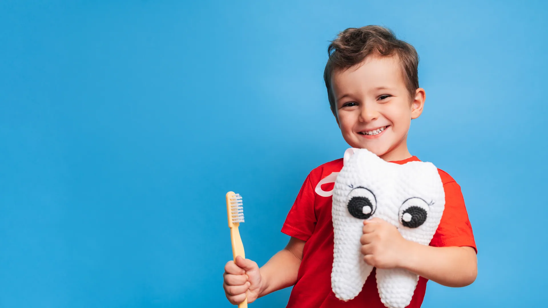 A happy young boy smiling while holding a toothbrush and a friendly plush tooth toy